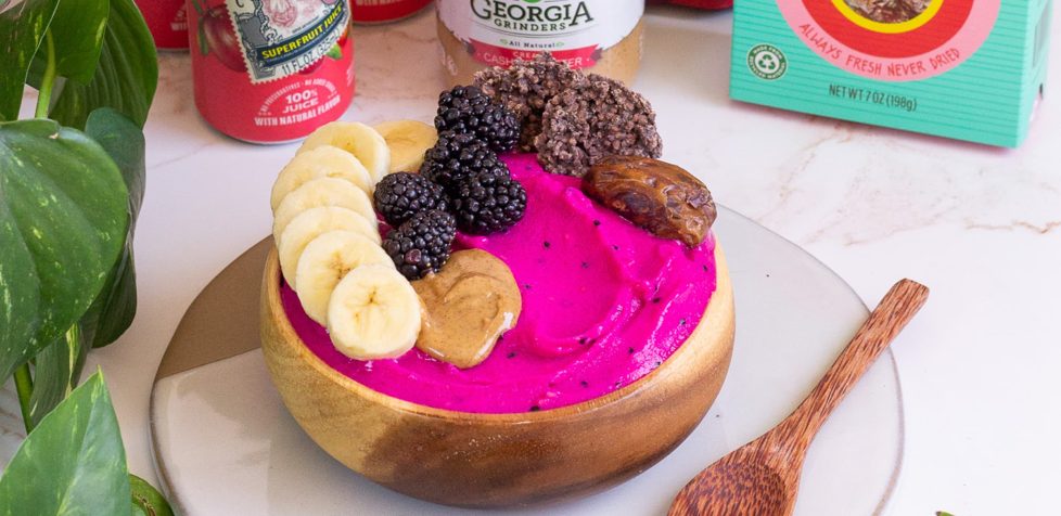 Pink mango smoothie bowl presented in a coconut bowl surrounded by ingredients and a wooden spoon